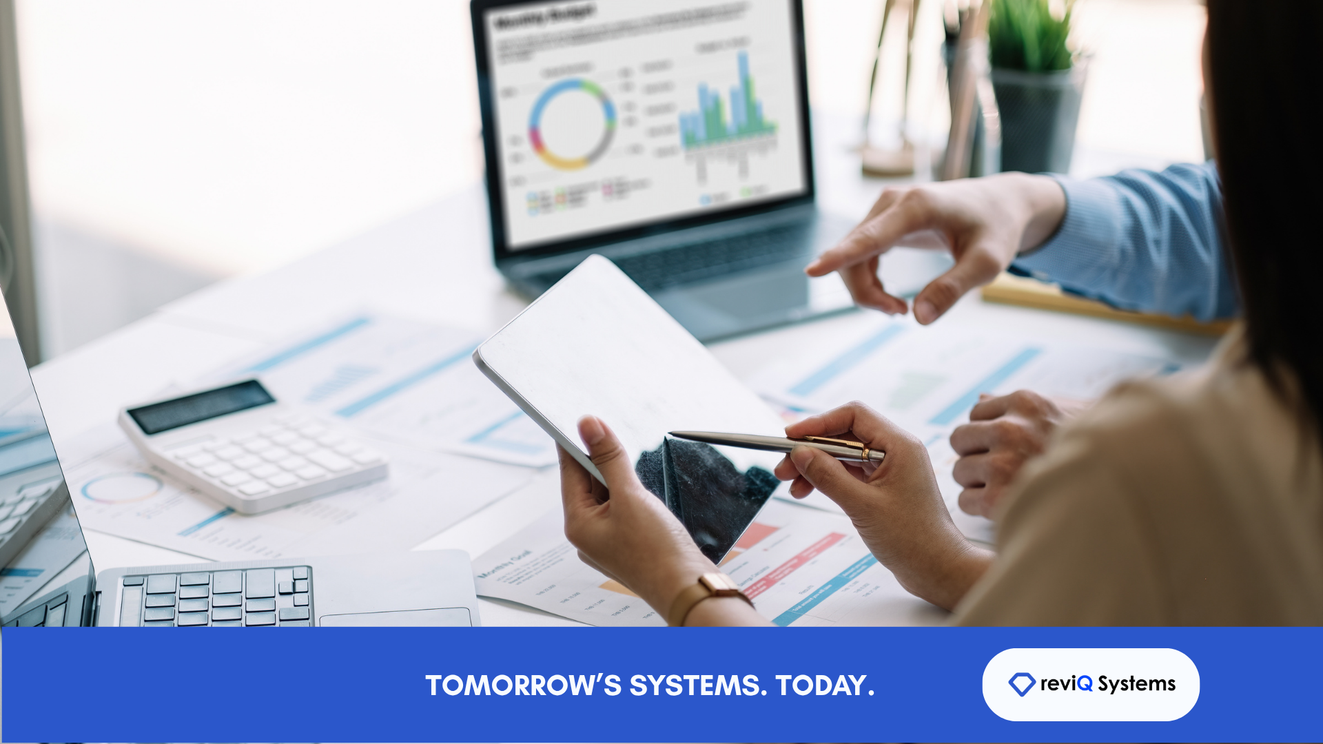 Two professionals collaborate over business performance data at a bright office desk. One holds a tablet and points with a pen while another gestures toward reports and a laptop displaying colourful charts and graphs. Printed analytics sheets, a calculator, and another laptop sit on the desk. A blue banner across the bottom reads “TOMORROW’S SYSTEMS. TODAY.” alongside the reviQ Systems logo.